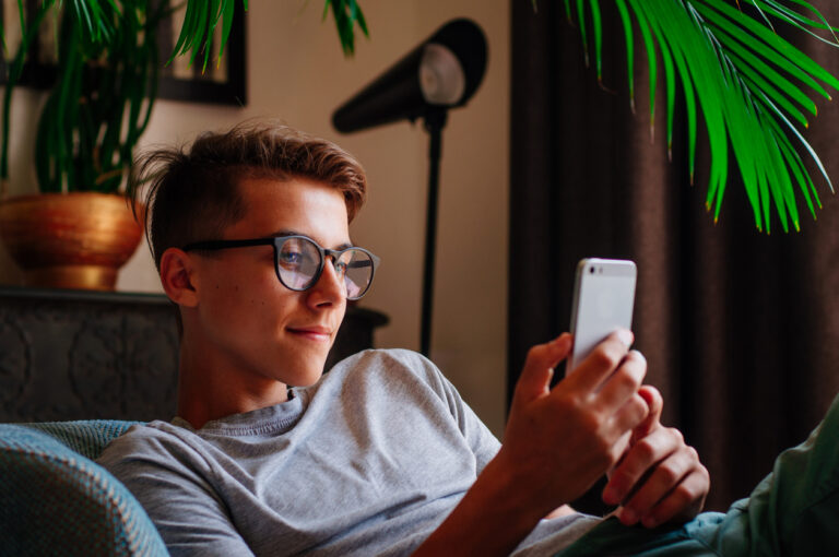 Generation Z concept. Smiling boy in glasses using smartphone sitting in the chair indoor.