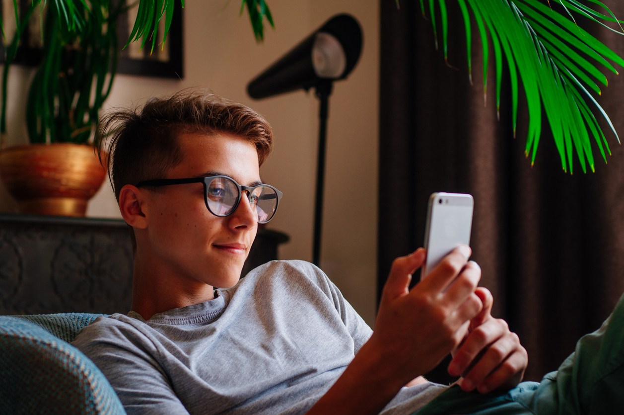 Generation Z concept. Smiling boy in glasses using smartphone sitting in the chair indoor.
